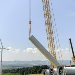 A crane lifting a section of a wind turbine during construction in a meadow on a sunny day.