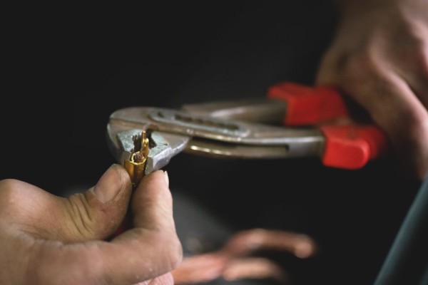 A man's work-worn hands using a pair of red-handled pliers to crimp a wire with a gold-colored connector.