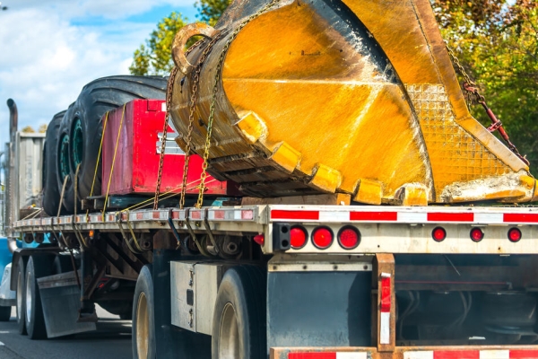 A semi-truck with a flatbed trailer is hauling large tires, a large red dumpster, and a yellow excavator scooper.