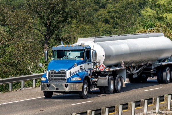 A blue semi-truck pulls a white tanker trailer down the road. The road has metal guardrails and trees lining it.