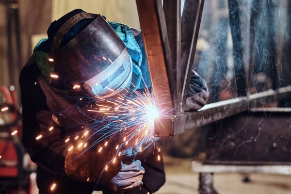 A person wearing a protective face mask, gloves, and jacket uses a tool to weld together two pieces of metal.