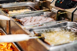 Four metal baskets filled with different kinds of food submerged in bubbling oil in deep fryers.