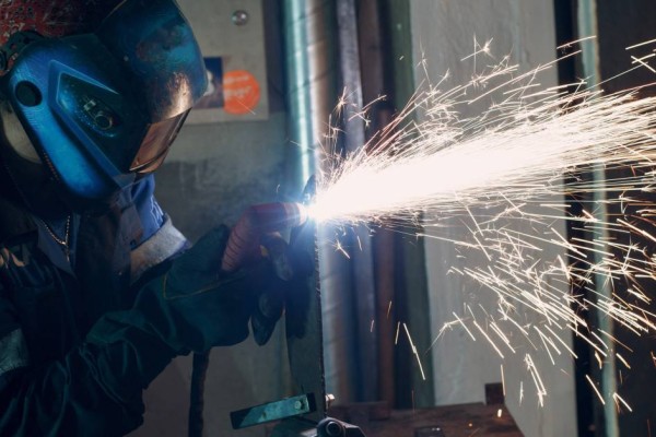 A man wearing a flame-resistant bodysuit and a face shield is using a plasma torch to cut a metal part in a workshop.