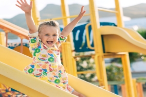 A little girl in a dress featuring cartoon fruit slices goes down a yellow playground slide, laughing and holding her arms up.