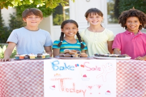 Four children are standing behind a table with cupcakes. The table has a sign that says "Bake Sale Today."