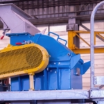 A yellow, blue, and silver hammer mill sits on a metal stand in the middle of an oversized, metal building.