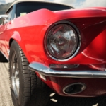 A classic muscle car, painted in shiny red, is parked on the side of the road with its hood and headlight in view.