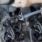 A close-up view shows a person using a silver wrench and a silver hex key to adjust a vehicle's engine.