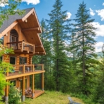 The exterior of a multistory log cabin with a deck on each level overlooking mountains and a forest.