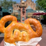 A person holds a mouse-ear-shaped pretzel in front of a theme park attraction entrance.