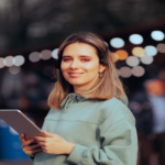 A smiling woman wearing a light-green sweatshirt and a black headset holds a silver tablet in her hands.