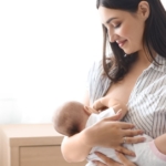A young woman in a striped short-sleeve shirt breastfeeds her newborn, cradling the infant in a bright room.