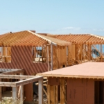 A partially built home stands near tall grass and weeds, with the ocean and a clear blue sky in the background.