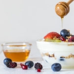 A small, glass bowl filled with yogurt is topped with granola and berries. A hand holds a honey wand and drizzles honey on top.