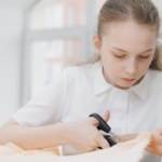 A girl in a white shirt in front of a window looks down and uses a pair of scissors to cut through a piece of fabric.