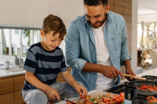 A parent cooking over a stovetop and leaning over to observe as their child cuts a variety of vegetables on a cutting board.