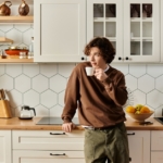 A person leans on a counter in their cozy and modern kitchen space while they drink out of a small cup.
