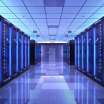 A vast data center with server racks on either side of a long hallway extending toward a door. They are casting a blue light.