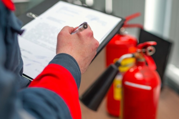 A close-up of an engineer professional wearing a dark blue uniform, inspecting a fire extinguisher with a clipboard.