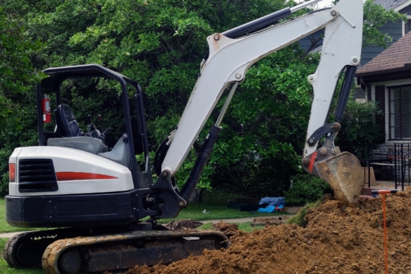 An unmanned white and red excavator in the front yard of a home with its arm next to a large pile of dirt.