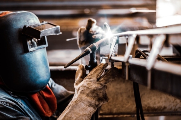 A person wearing a welding helmet, leather jacket, and gloves using a torch to weld metal in a workshop.