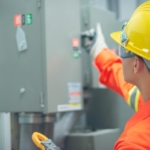 An engineer wearing protective equipment holds a multimeter with one hand and opens a control cabinet with the other.