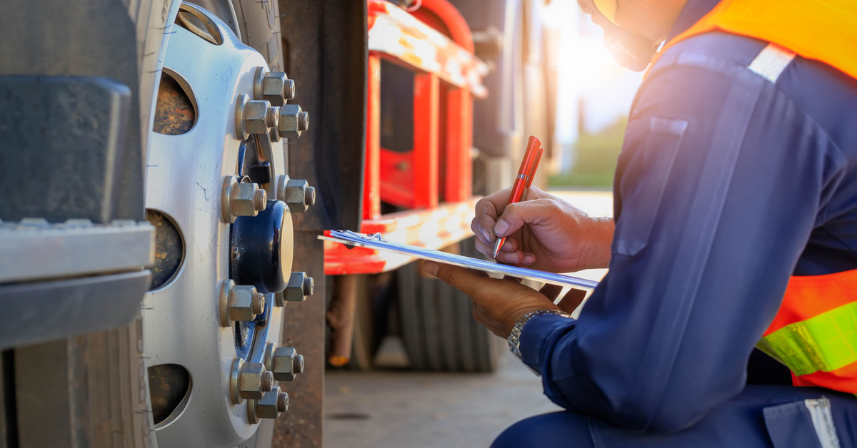 A worker in a safety vest and hard hat inspects a truck wheel, writing on a clipboard under bright sunlight.