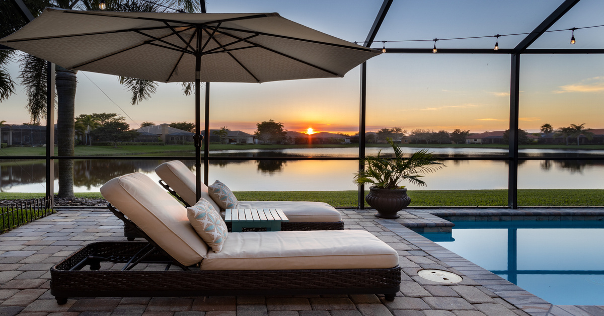 Two lounge chairs with tan cushions sit on a brick patio next to an in-ground pool in a residential backyard.