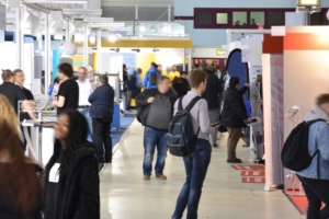 Various groups of people visiting at and walking between different booths at a trade show. The event floor is brightly lit.