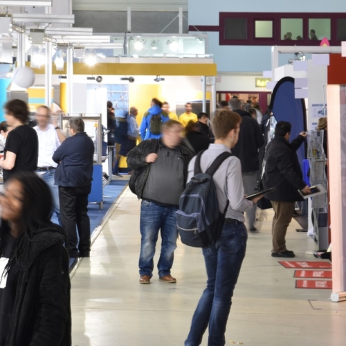 Various groups of people visiting at and walking between different booths at a trade show. The event floor is brightly lit.