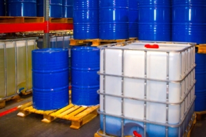 A clear, plastic, square container is surrounded by several blue barrels. The square container contains a blue liquid.