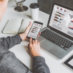 A woman sits at a desk with a laptop, phone, and tablet in front of her. There is also a blank, open notebook on the desk.