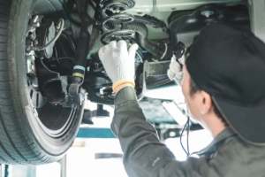 A car mechanic wearing gloves and a backwards hat works under a lifted vehicle. He touches the car's spring.