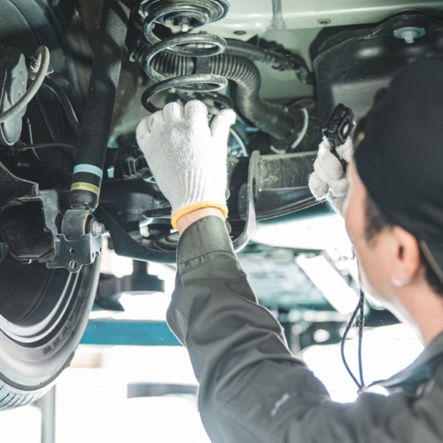 A car mechanic wearing gloves and a backwards hat works under a lifted vehicle. He touches the car's spring.