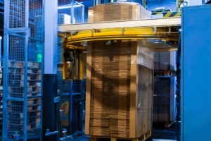 A pallet with a tall stack of corrugated cardboard folded into a tower inside the cage of a stretch wrapping machine.