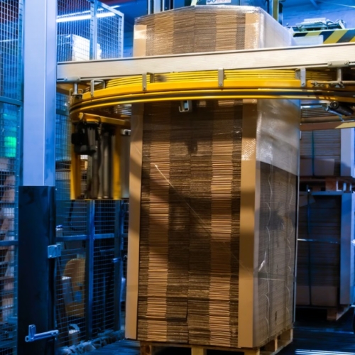 A pallet with a tall stack of corrugated cardboard folded into a tower inside the cage of a stretch wrapping machine.