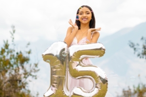 A teenage girl wearing a pink dress smiles while bubbles float around her. In front is a balloon that spells "15."