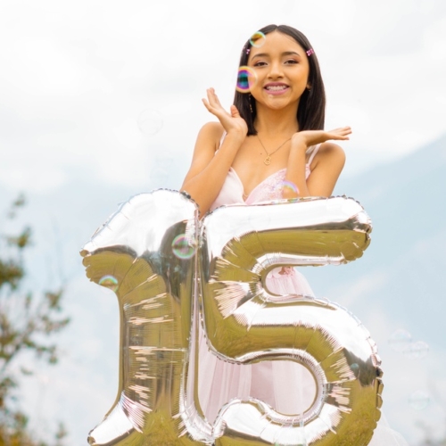 A teenage girl wearing a pink dress smiles while bubbles float around her. In front is a balloon that spells "15."
