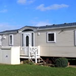 A mobile home with tan-colored siding has a small porch with white fencing. A white shed is in front of the porch.