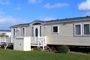 A mobile home with tan-colored siding has a small porch with white fencing. A white shed is in front of the porch.