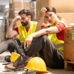Two stressed warehouse workers sitting next to boxes. They both have hard hats lying on the ground next to them.