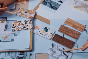 The hands of two different people point to color samples lying on home interior design plans. Both hands hold pencils.