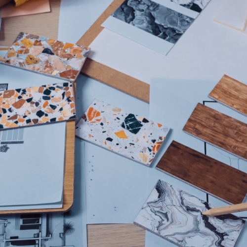 The hands of two different people point to color samples lying on home interior design plans. Both hands hold pencils.