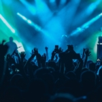 A band plays a concert onstage in front of a large crowd. The venue features blue and green lighting.