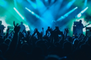 A band plays a concert onstage in front of a large crowd. The venue features blue and green lighting.