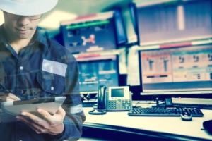 A man in blue coveralls and a white hard hat using a stylus on a tablet while standing near several monitors in a control room.