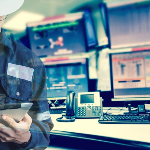 A man in blue coveralls and a white hard hat using a stylus on a tablet while standing near several monitors in a control room.