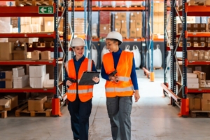 Two workers in safety gear are checking inventory on a clipboard in a large warehouse with packages on each shelf.