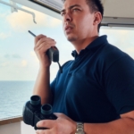 A man wearing a blue polo shirt, holding a radio with one hand and binoculars with the other, inside a boat cabin.