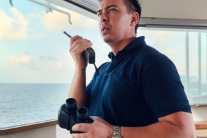 A man wearing a blue polo shirt, holding a radio with one hand and binoculars with the other, inside a boat cabin.